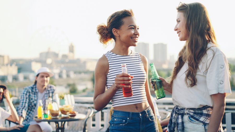 Zwei junge Frauen lachen mit Getränken auf einer Dachterrasse